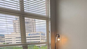 A room with a brown sofa, a small black table with a plant, a floor lamp, and a window with blinds overlooking a building. - Deep Ellum-Sleeps 5-Balcony-Pool-Medical District (Dallas)