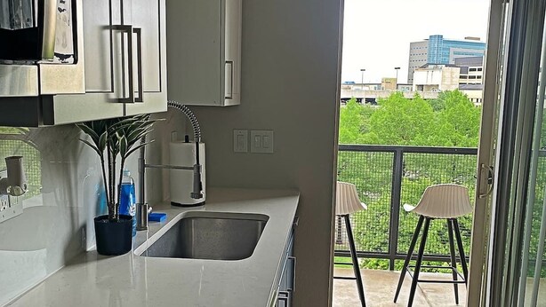 A modern kitchen with a built-in stove, a sink, and cabinets. There is a balcony with a chair and a table, and a view of greenery and buildings outside. - Deep Ellum-Sleeps 5-Balcony-Pool-Medical District (Dallas)