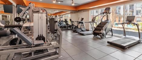Un gimnasio con diferentes máquinas de ejercicio, ventanas amplias y piso de baldosas.
