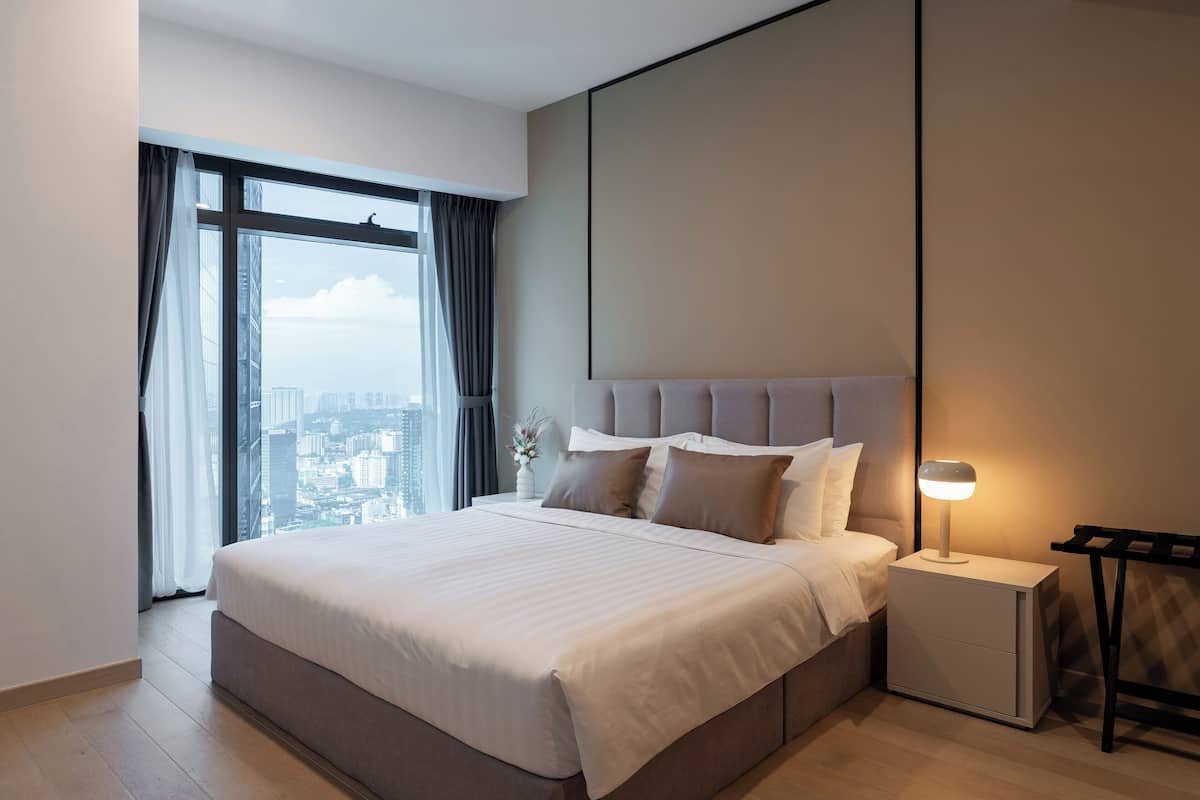 Habitación de hotel moderna con una cama grande, una mesita de noche con lámpara y vistas a la ciudad a través de amplios ventanales.