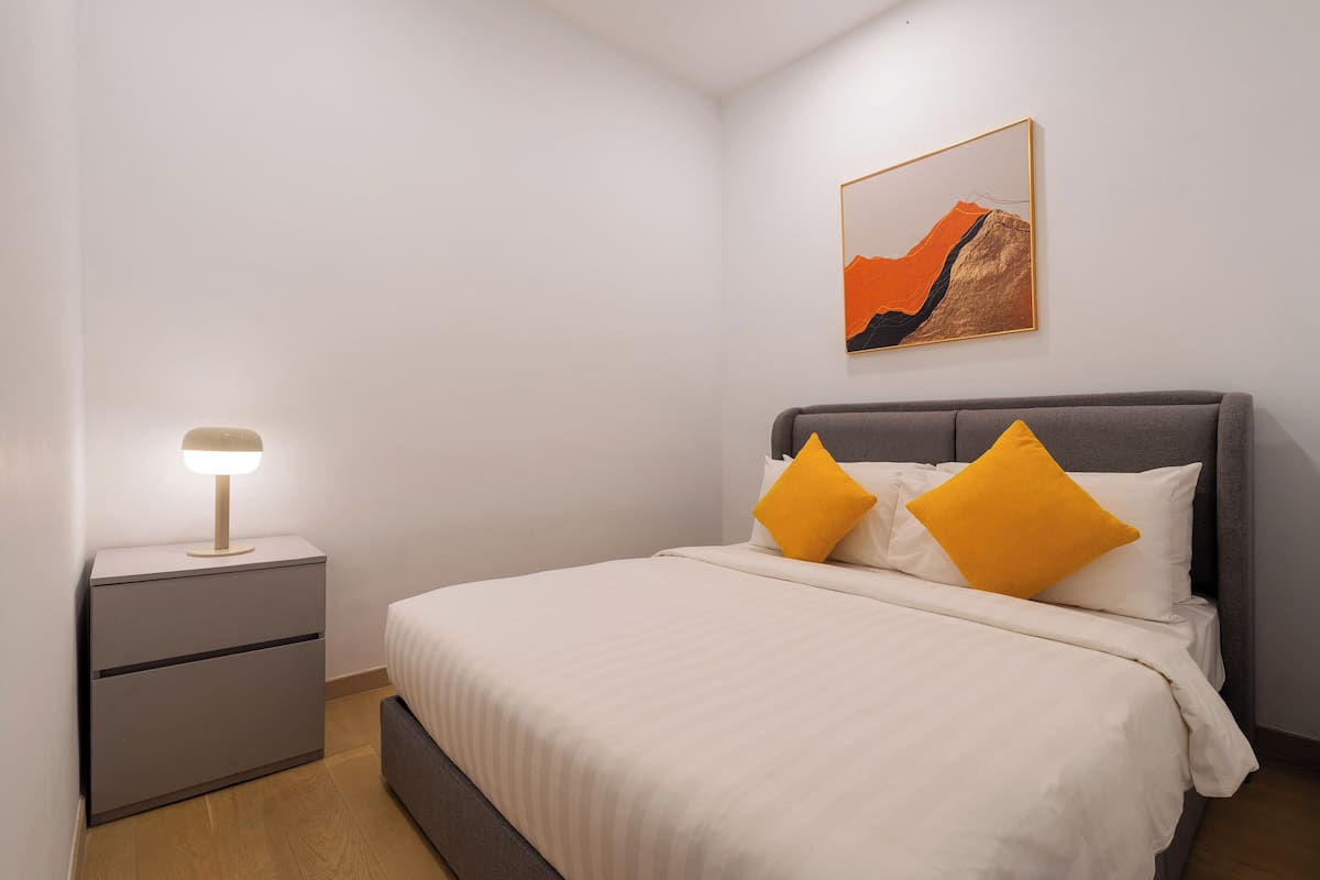 Una cama bien hecha con dos almohadas, una mesita de noche con una lámpara, y un cuadro en la pared.