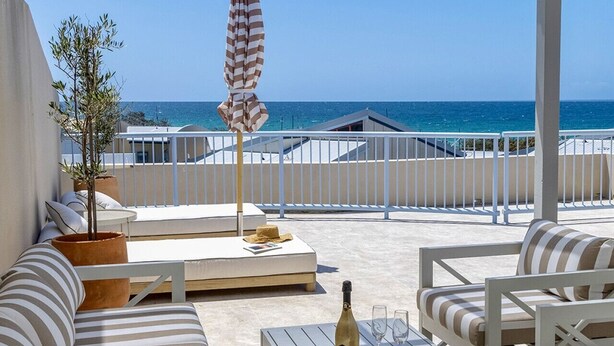 Un espacio de estar al aire libre cubierto con mobiliario blanco, una mesa con una botella y vasos, y vistas al mar.