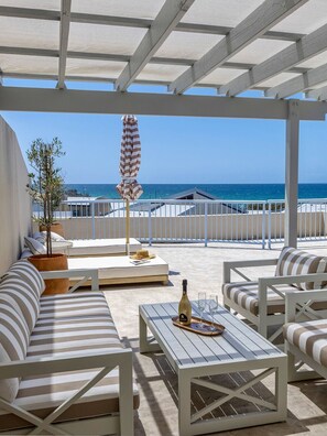 A covered outdoor seating area with white furniture, a table with a bottle and glasses, and a view of the sea.