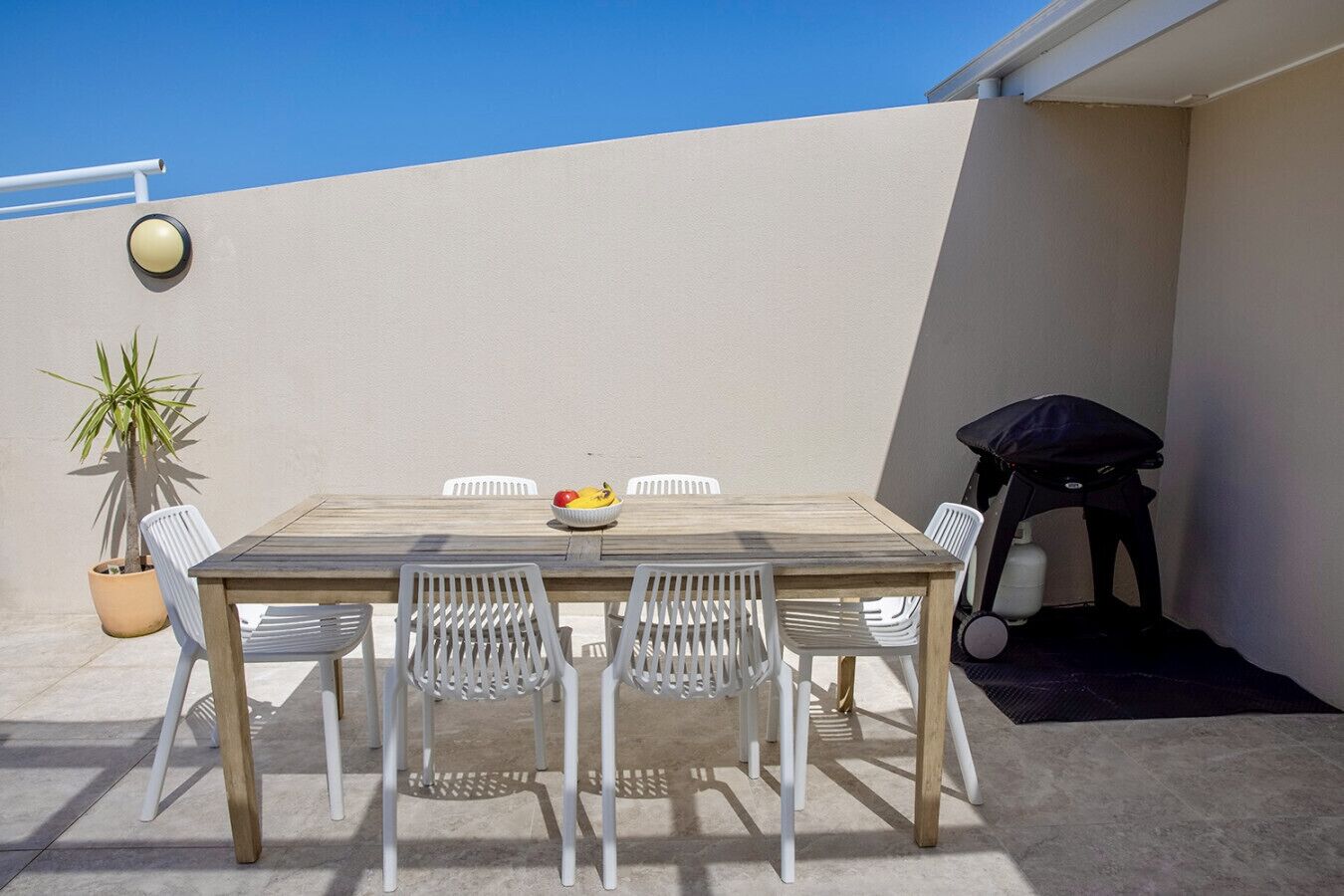 An outdoor patio with a wooden table and white chairs, a potted plant, and a barbecue grill.