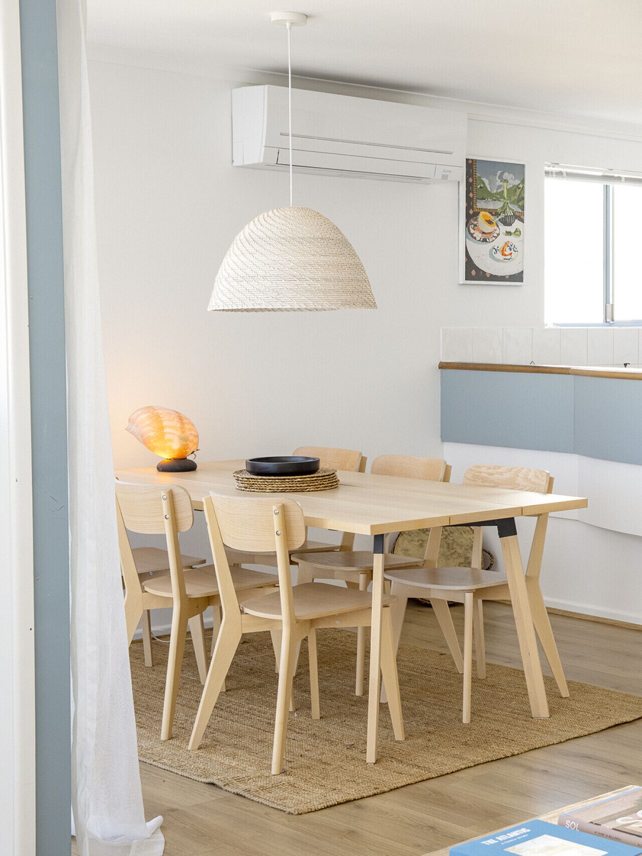 A dining area with a wooden table and chairs, a woven pendant light, and a wall-mounted air conditioning unit.