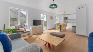 Een moderne woonkamer met een bank, een salontafel en een televisie. Er is een keukenhoek met witte kasten en een eettafel met kleurrijke stoelen.