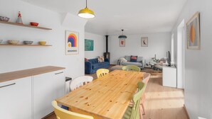 Een moderne keuken met houten kastjes, een eettafel en kleurrijke stoelen. Er staan planken met kommen en er hangt een lamp.