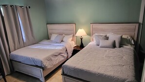 Duas camas de solteiro com cabeceiras, uma mesa de cabeceira com abajur e uma janela com cortinas.