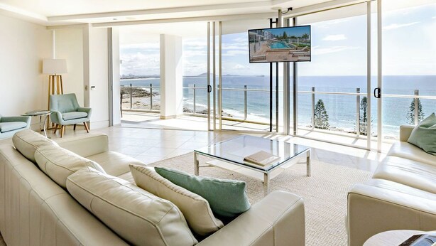A spacious living room with a large sofa, a coffee table, and a TV mounted on a glass wall.