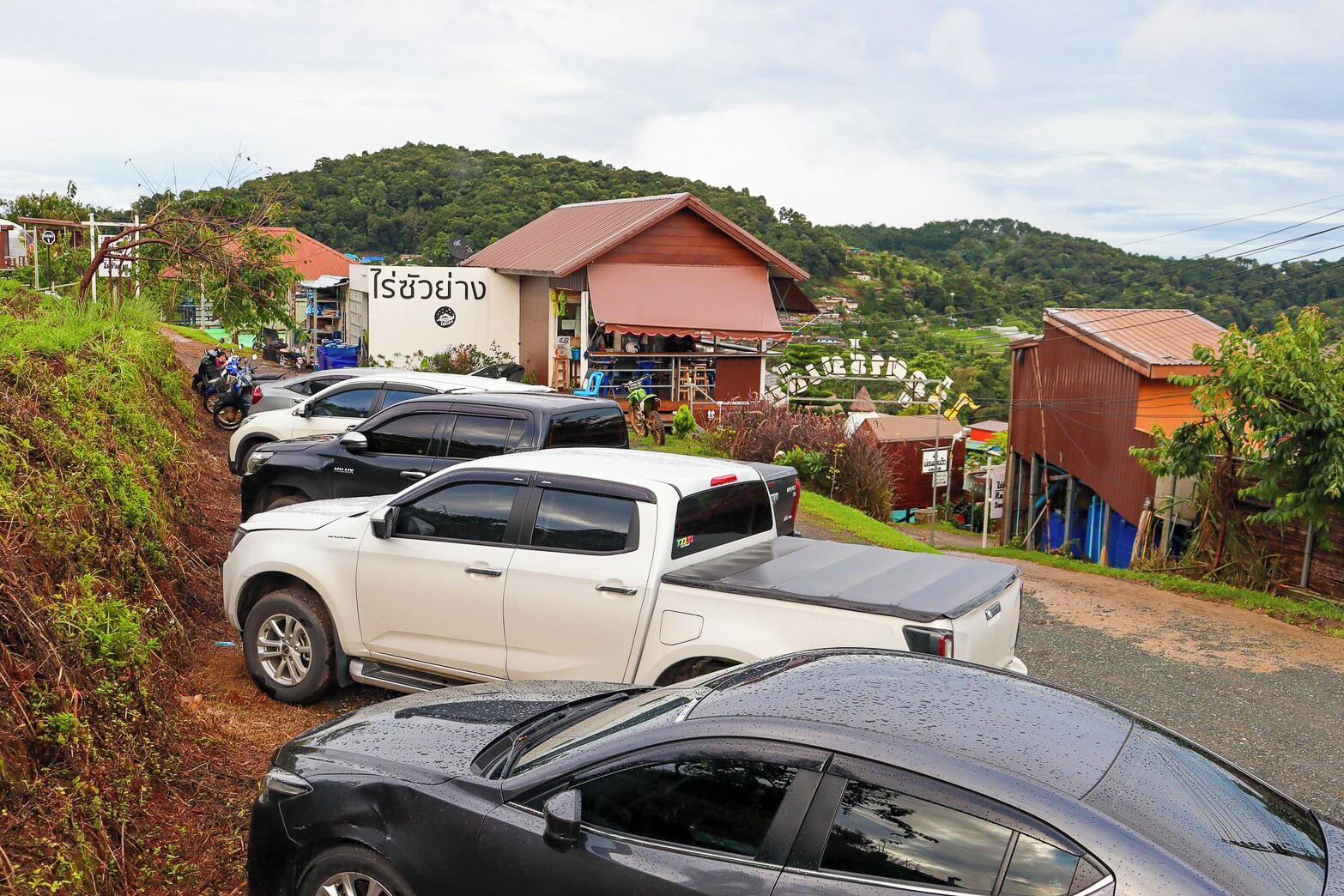 A parking area with multiple vehicles, a building with a sign, and a hilly landscape in the background.