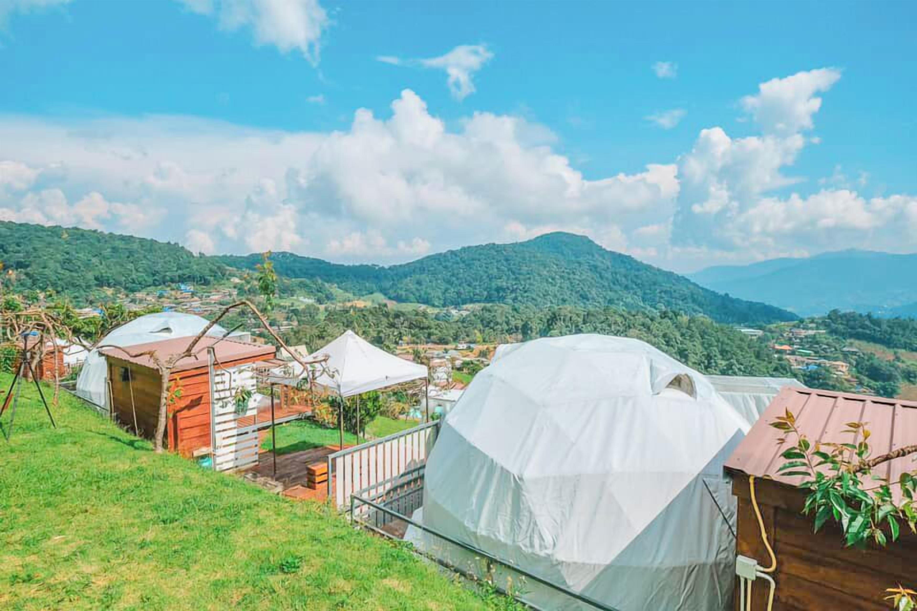 Vista de una colina con varias tiendas en forma de cúpula y una cabaña de madera, rodeado de abundante vegetación y montañas a la distancia.