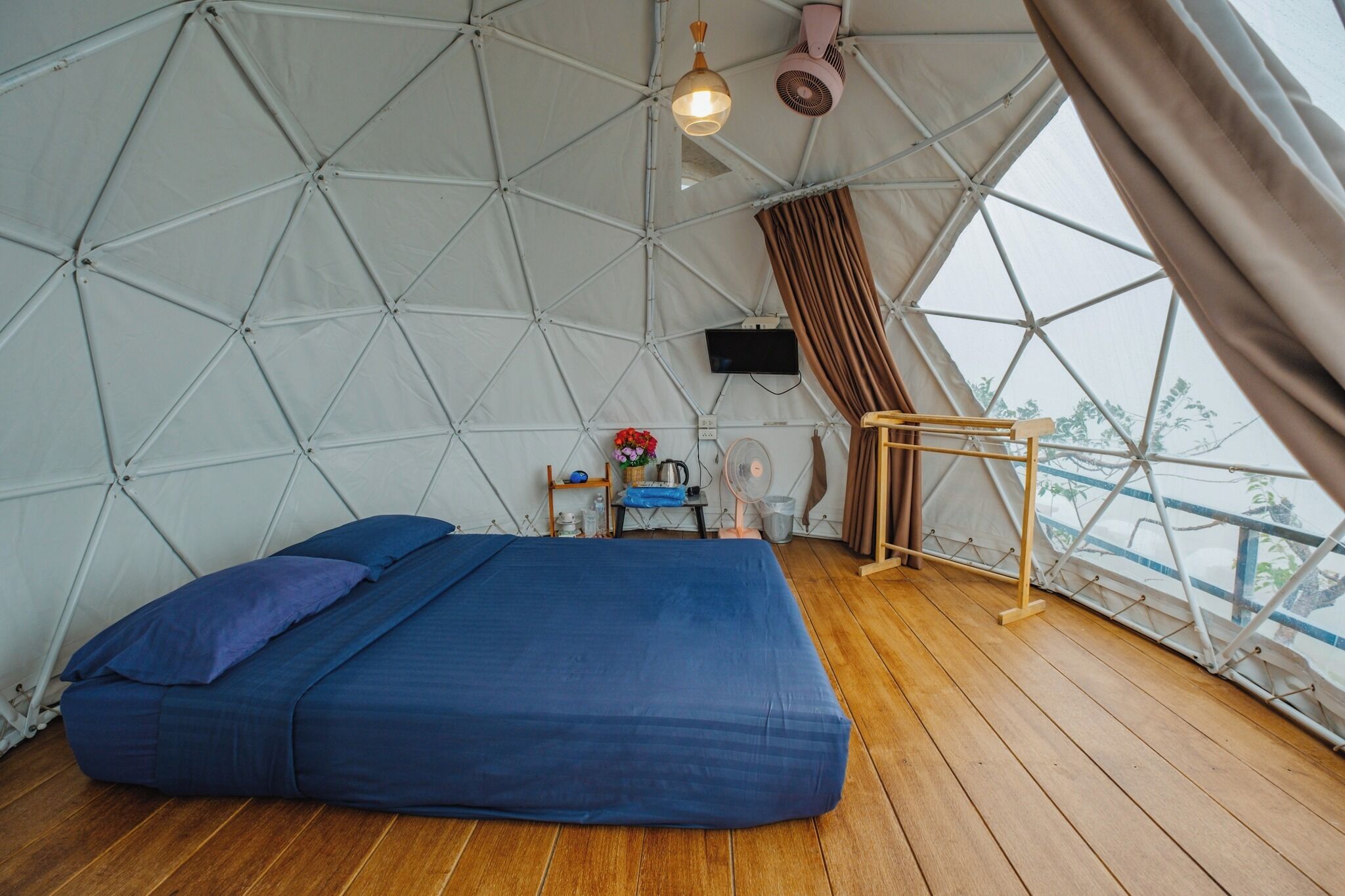 A tented room with a bed, wooden flooring, a small table with a kettle, and a view through the curtain.
