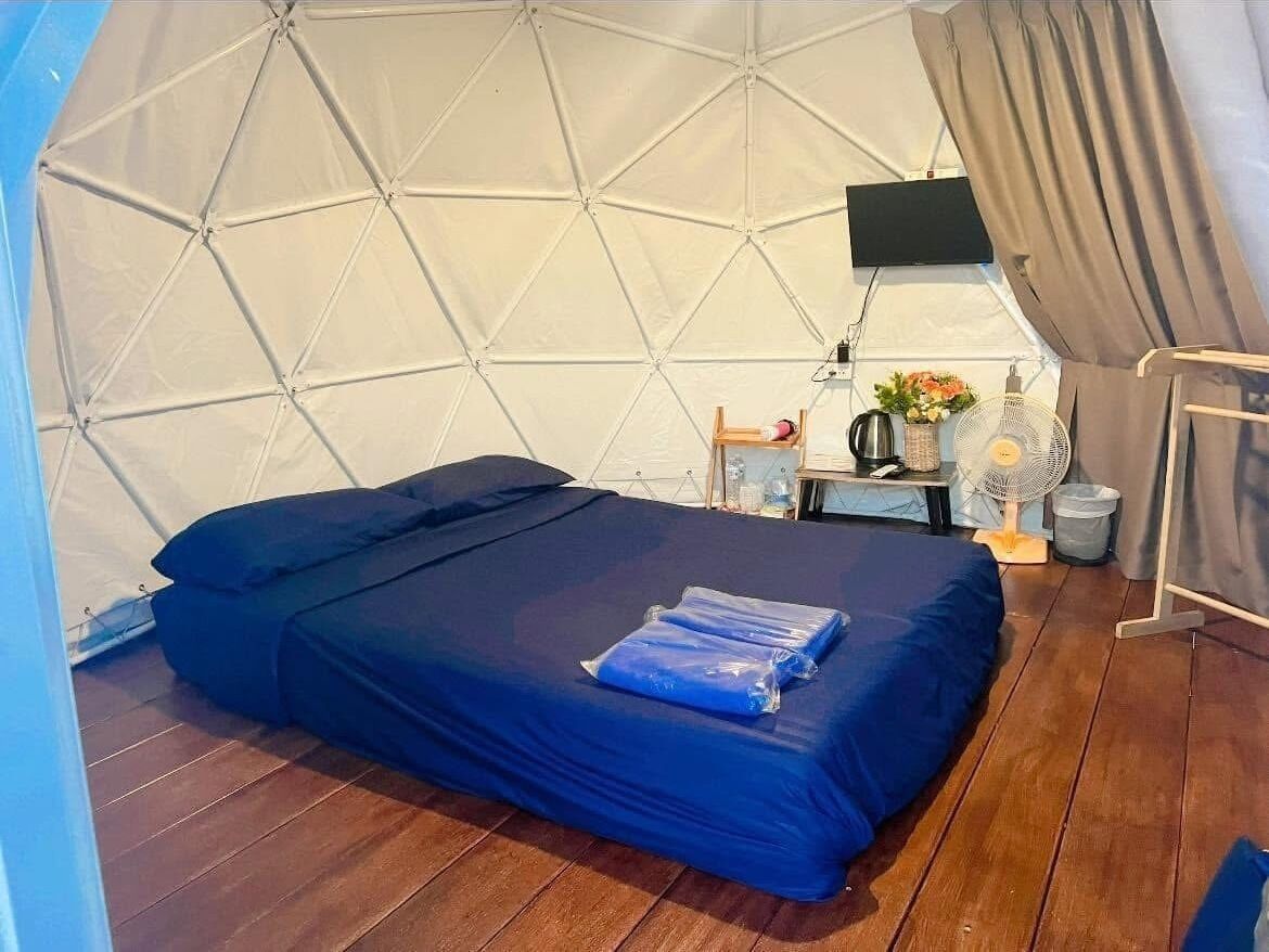 A tent-style room with a bed, a flat-screen TV, a small table with a fan, and a window with curtains.
