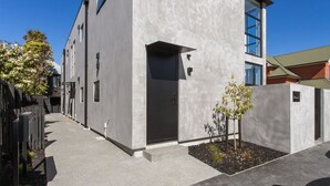 Un bâtiment moderne avec une façade en béton, une grande entrée et un petit espace jardin agrémenté d’un arbre.