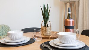Une table à manger dressée avec des bols, des assiettes et des couverts blancs, un panier tressé contenant une plante, une bouteille de couleur cuivrée et une lampe en arrière-plan.