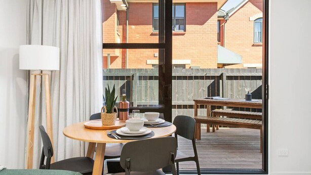 Un coin repas avec une table dressée pour deux personnes, une plante en pot et une vue sur un bâtiment en briques, visible à travers une porte vitrée.