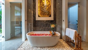Un baño moderno con una bañera independiente llena de pétalos de rosa, una obra de arte grande enmarcadas arriba de la bañera, y una silla de madera con una toalla.