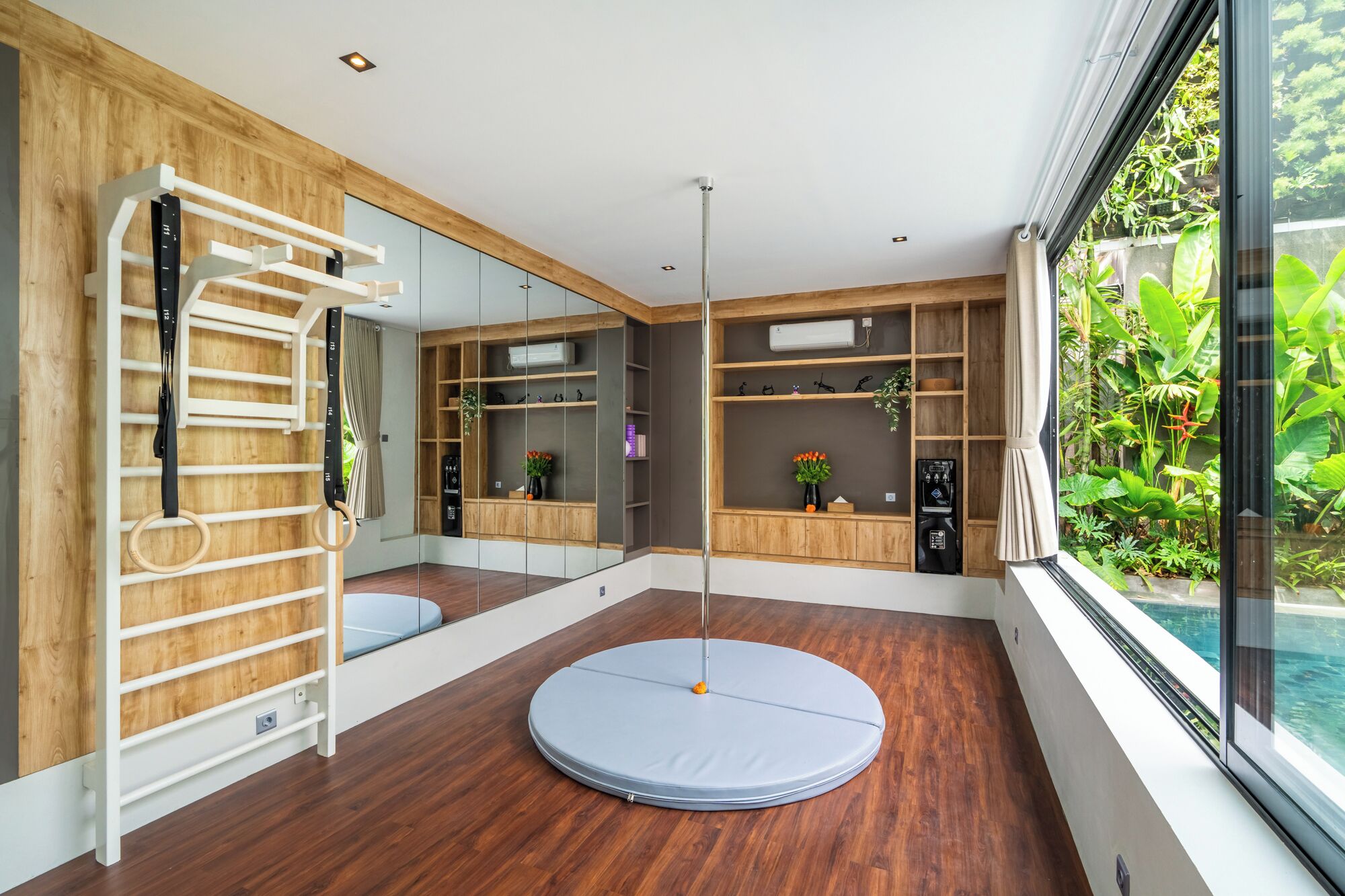 A modern room with a wooden floor, a pull-up bar, a ceiling fan, and a large window with a view of greenery.