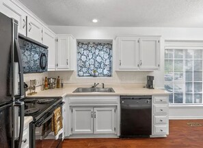 A kitchen with white cabinets, a stainless steel refrigerator, a dishwasher, and a window with patterned curtains.