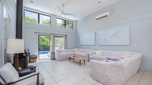 Una sala de estar amplia con un sofá modular grande, una mesa de centro de madera y una lámpara. Hay dos ventanas que dejan entrar luz natural y un ventilador de techo.
