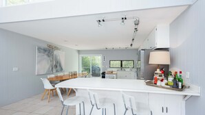 Una cocina moderna con armarios blancos, una isla central con taburetes, un comedor con mesa y sillas de madera, y un cuadro abstracto grande en la pared.