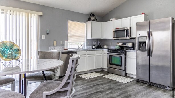A modern kitchen with white cabinets, stainless steel appliances, a dining area with a round table and chairs, and a window with blinds.
