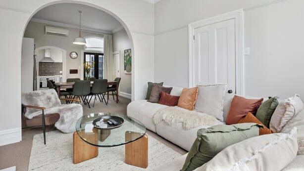 A modern living room with a glass coffee table, a sofa with multiple cushions, and a dining area in the background. - Villa on Rattray (Dunedin)