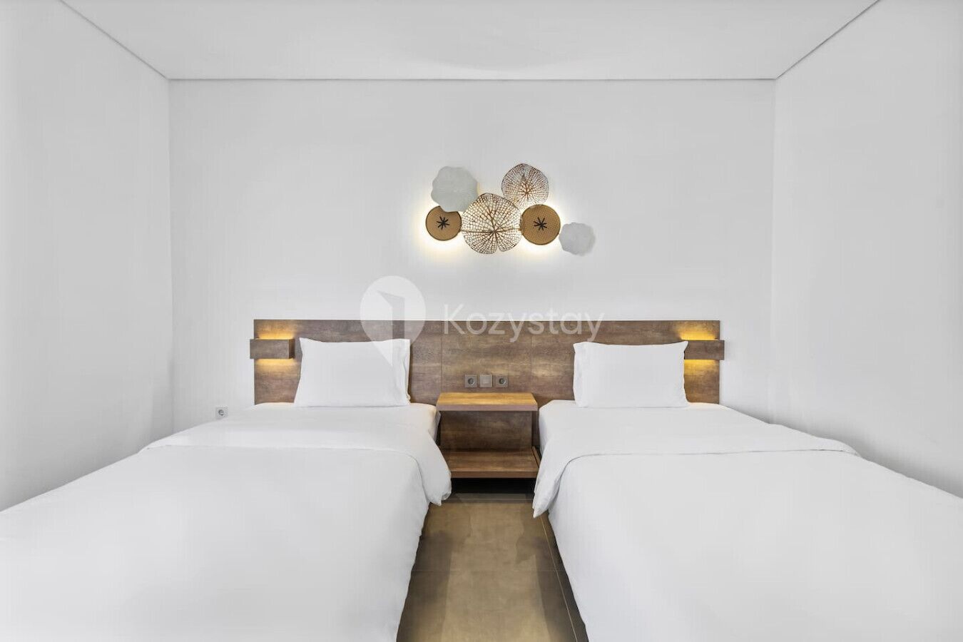 A modern hotel room with two single beds, a wooden headboard, and a decorative wall light fixture.