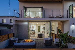 A modern two-story house with a spacious outdoor patio featuring a fire pit, seating, and a view into the interior.