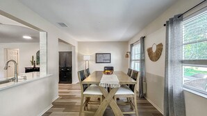 Ein Essbereich mit Holztisch, Stühlen und einer Obstschale. An der Wand hängt ein Spiegel, eine Tür führt in einen weiteren Raum, und ein Fenster ist mit Vorhängen drapiert.