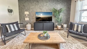Ein Wohnzimmer mit einem großen Flachbildfernseher, zwei schwarzen Sofas mit gestreiften Kissen, einem Holztisch und einer großen Kübelpflanze.