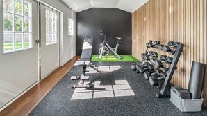 Ein Fitnessraum mit Trainingsgeräten, einem Ergometer und einem Wandregal für Kurzhanteln.