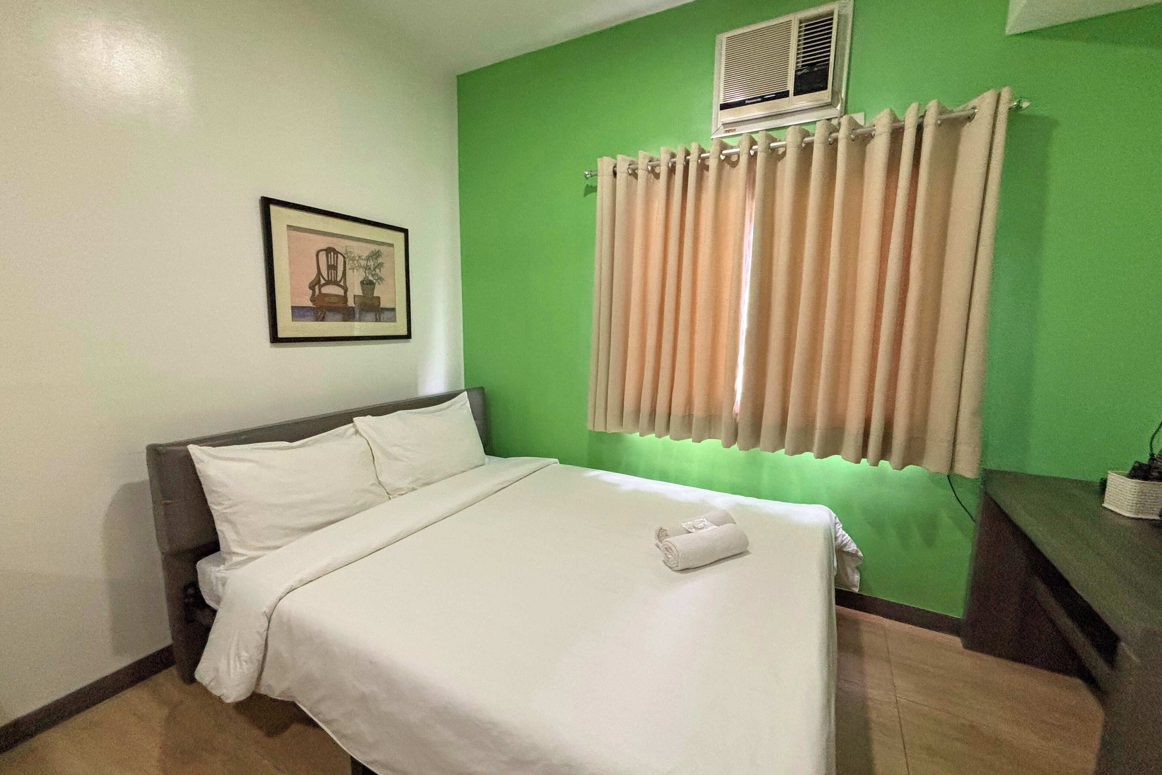 A neatly made bed with white linens, a framed picture on the wall, and a window with beige curtains.