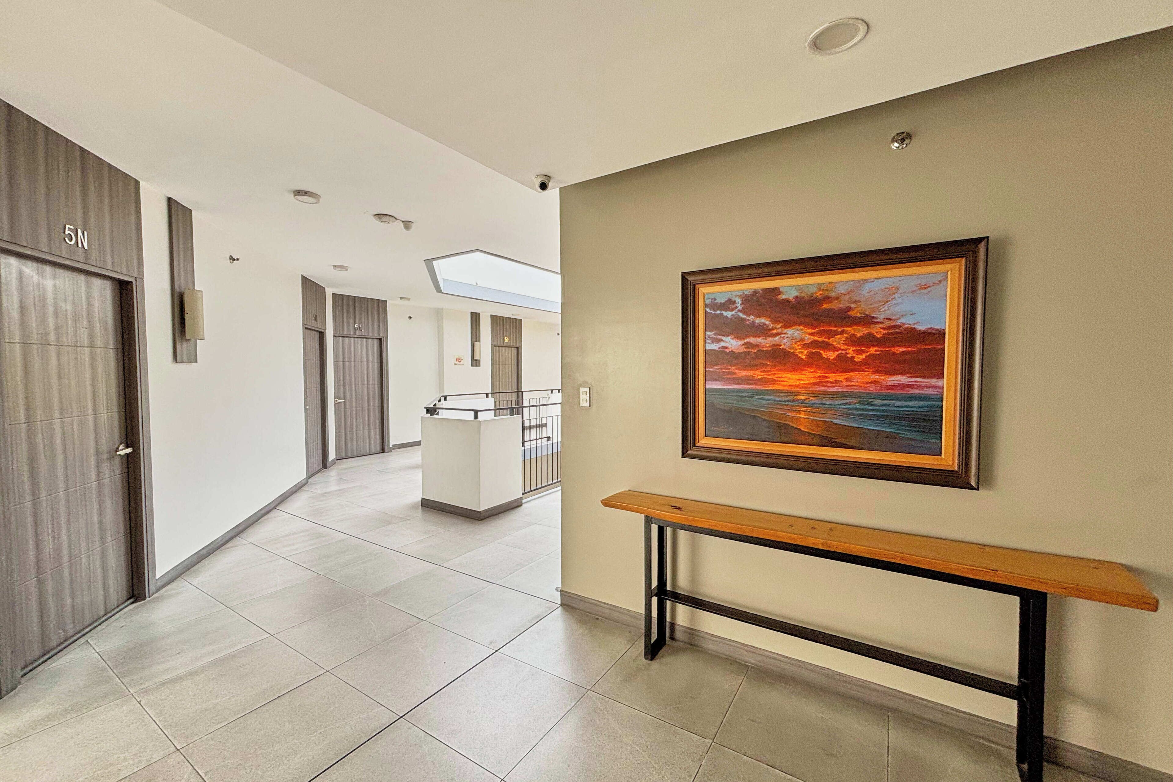 A modern hallway with a bench, a framed painting of a sunset, and a reception desk in the distance.