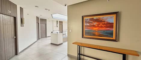 A modern hallway with a bench, a framed painting of a sunset, and a reception desk in the distance.