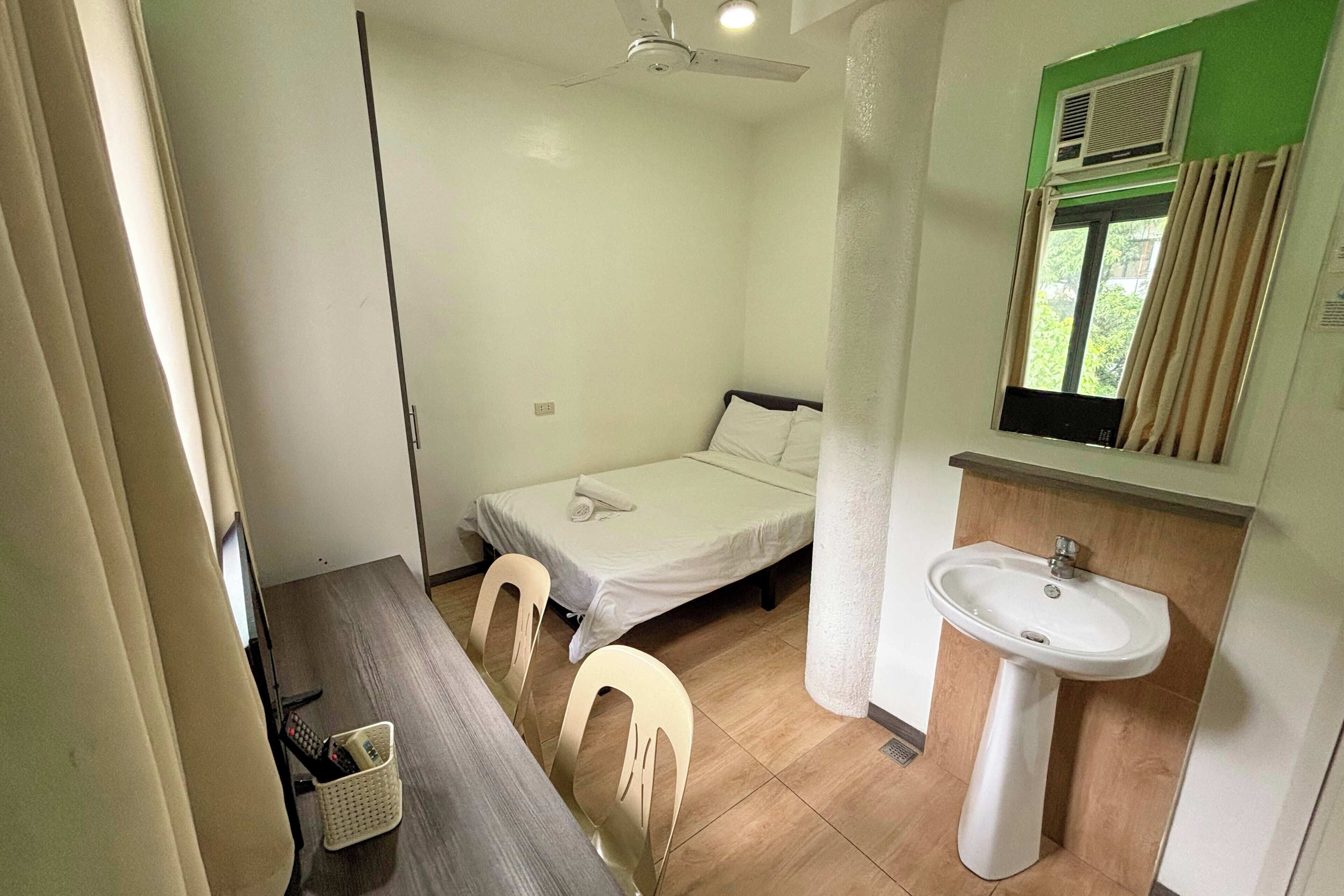 Habitació petita i neta amb un llit, una escriptori amb cadires, un rentamans i una finestra amb cortines.