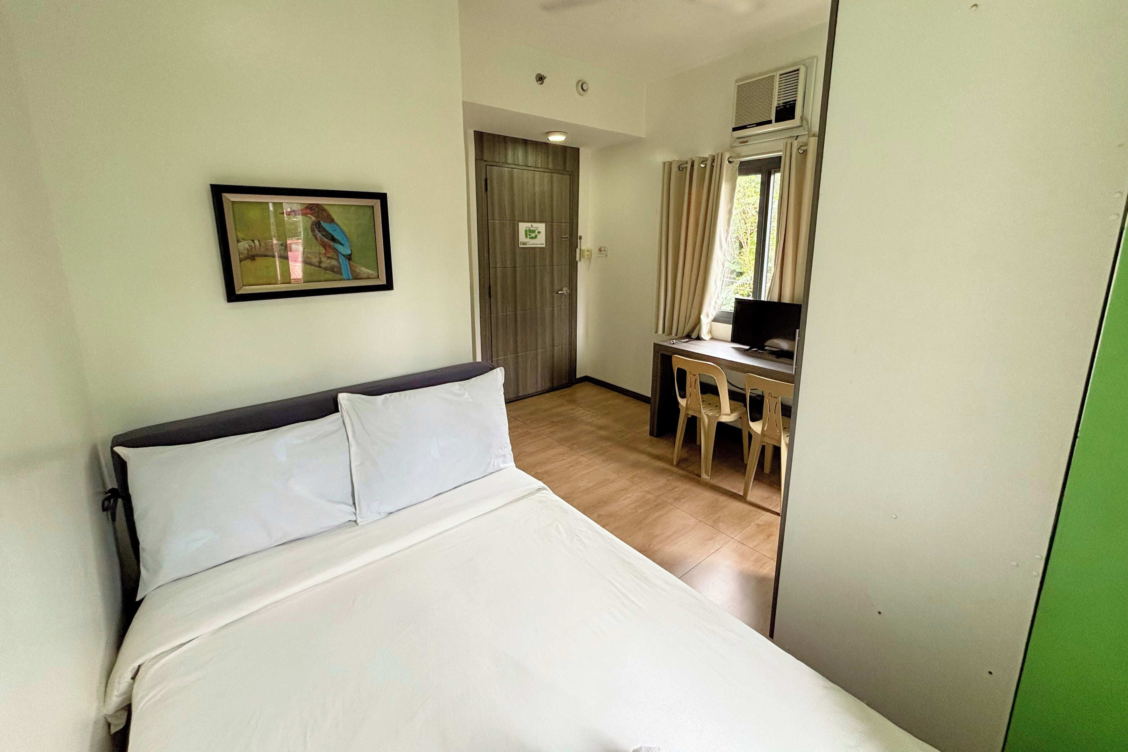 A hotel room with a bed, a desk with a chair, a window with curtains, and a framed picture on the wall.