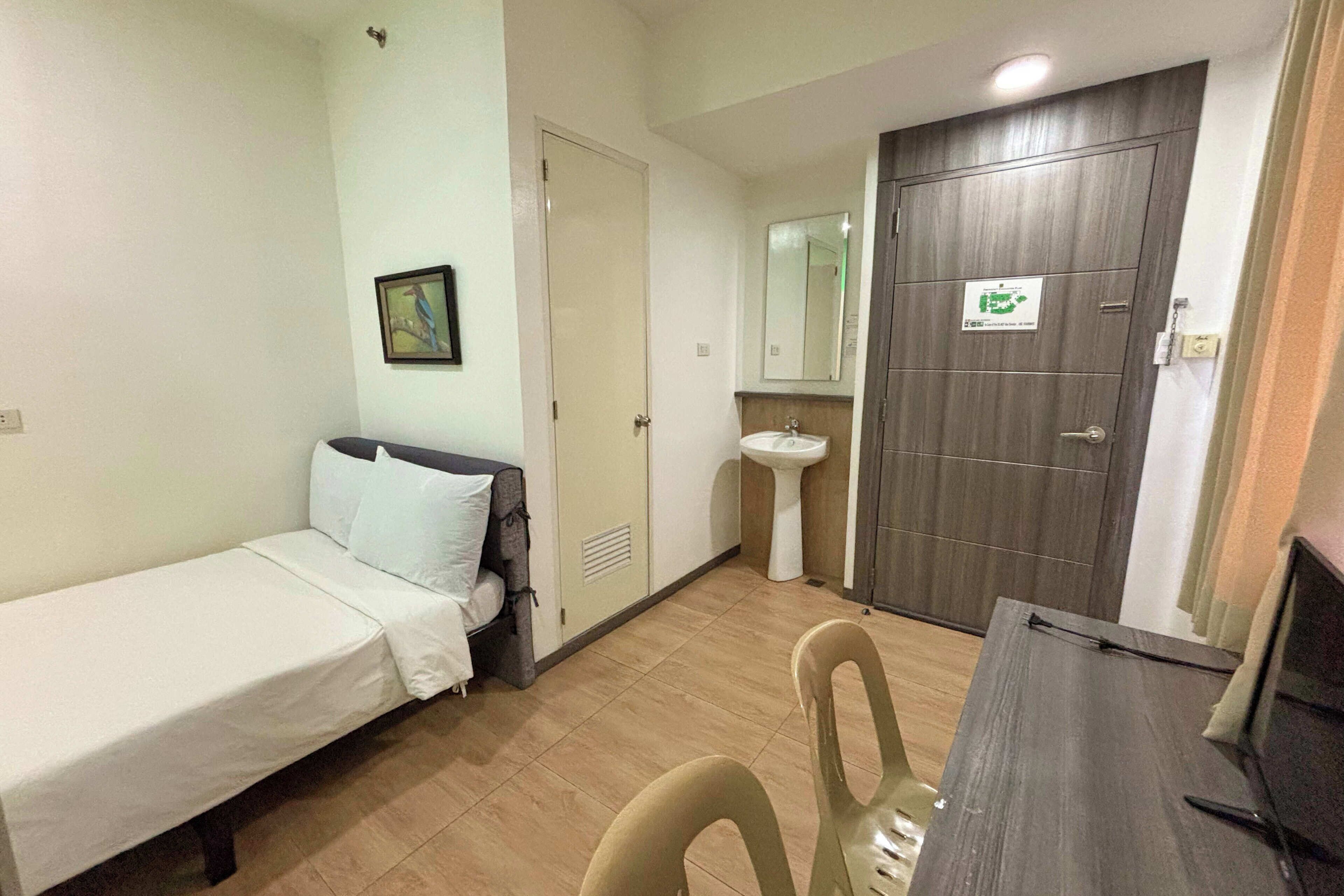 A small, clean room with a single bed, a bedside table, a chair, a desk, a sink, and a door with a mirror.