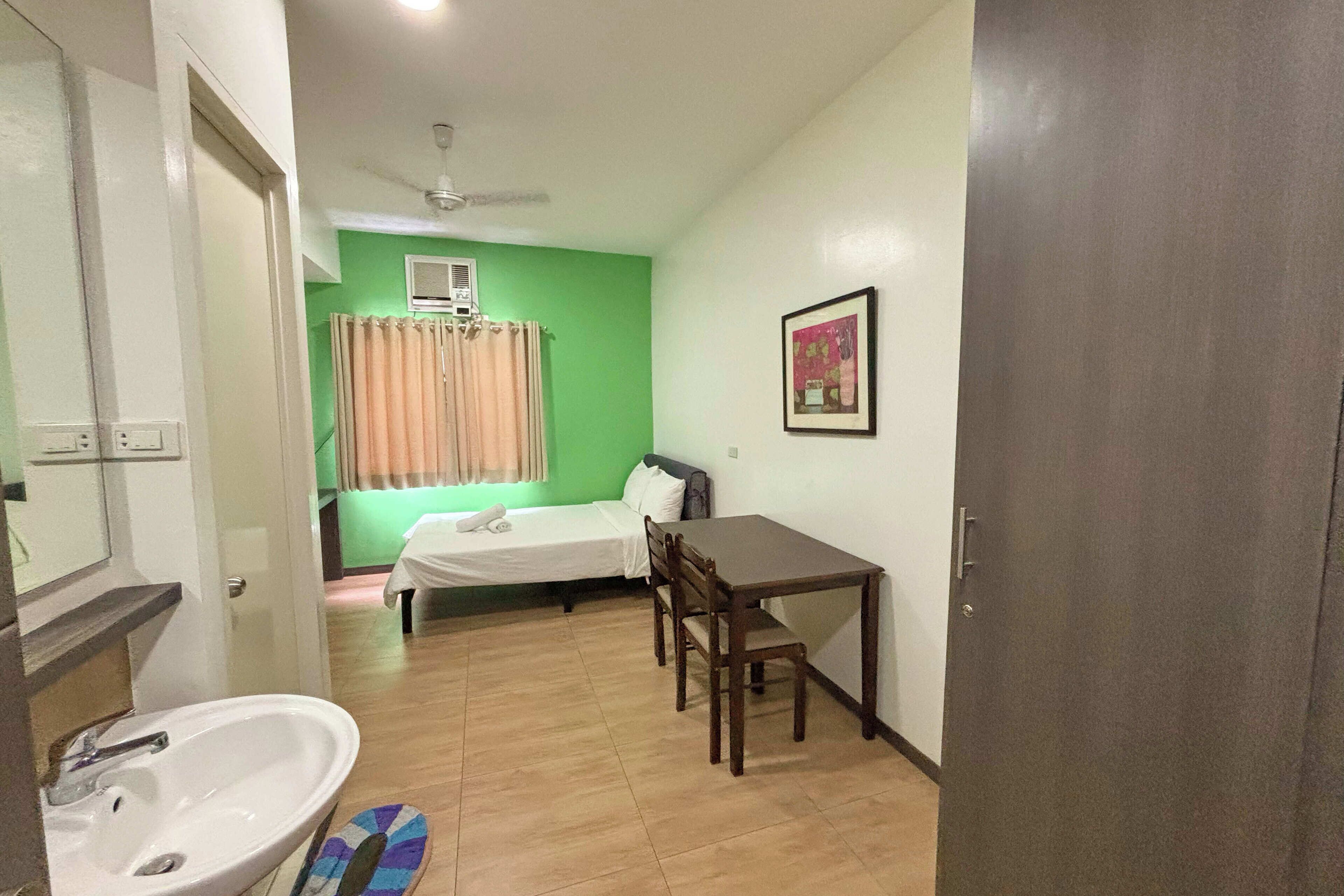 A small room with a bed, a table, a chair, a sink, and a ceiling fan.