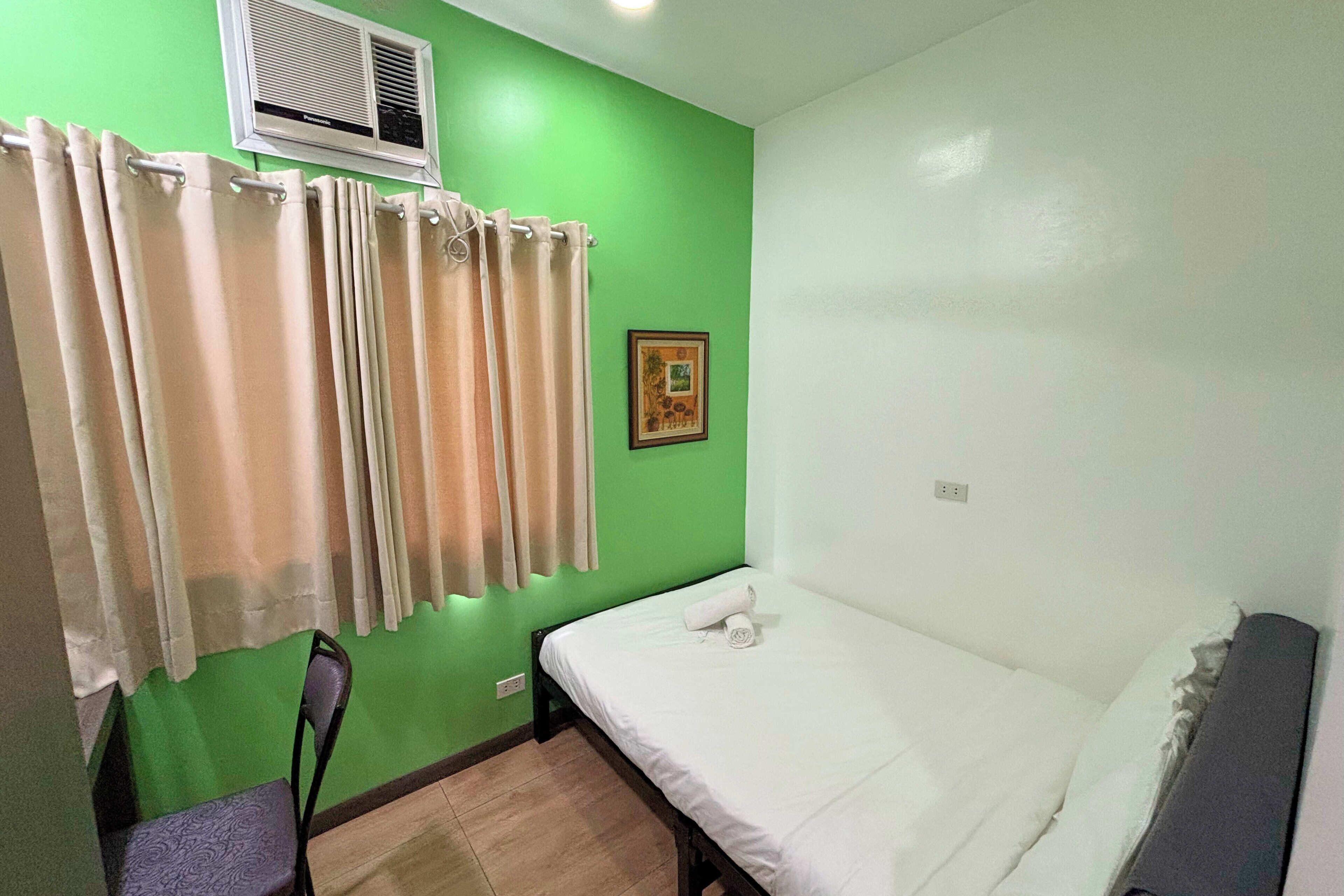 Une petite chambre avec un lit simple, un mur vert, une fenêtre avec des rideaux beiges, une chaise et un tableau encadré.