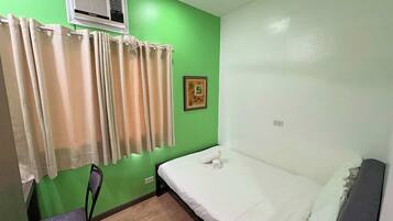 Una habitación pequeña con una cama individual, pared verde, ventana con cortinas beige, una silla e imagen enmarcada.