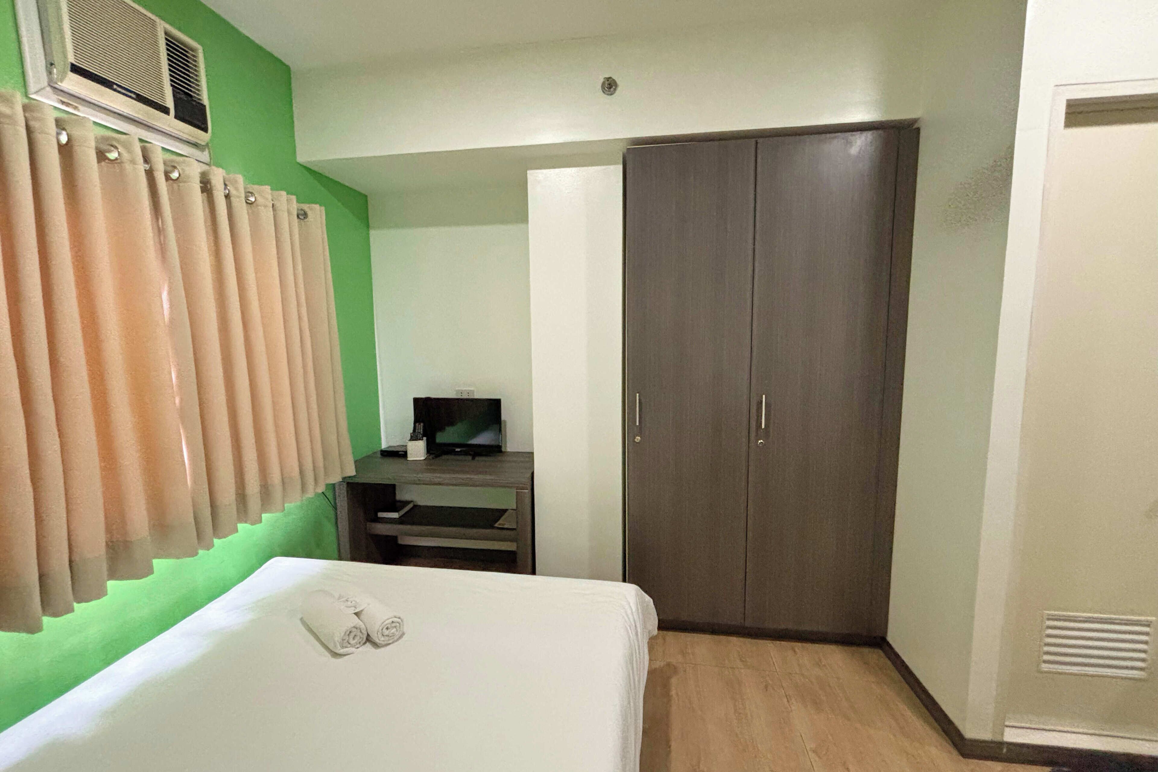 A hotel room with a bed, a wardrobe, a television on a stand, and a window with curtains.