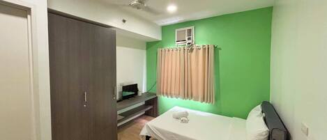 A small room with a bed, a wardrobe, a desk with a computer, and a window with curtains.