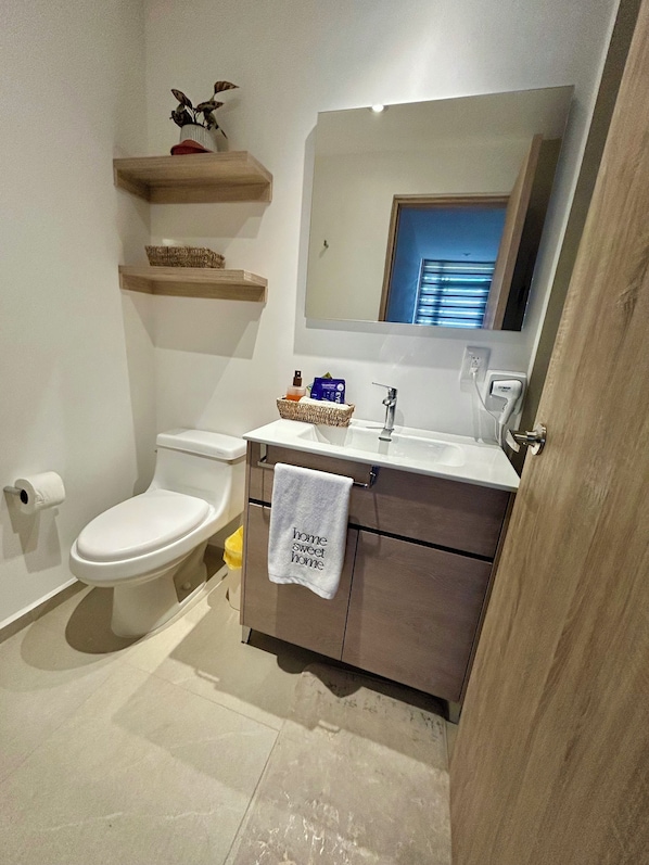 A bathroom with a toilet, sink, and mirror. There are wooden shelves above the toilet and a towel hanging on the door. - New modern apartment very well located! (Pocitos)