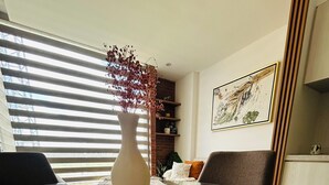 Een moderne woonkamer met een glazen salontafel, een beige vaas met gedroogde bloemen, een kamerkleed met dessin, een bank met kussens, en een kunstwerk in een lijst aan de muur.