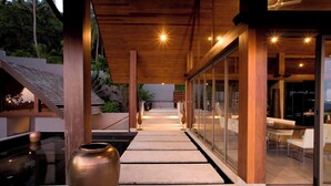Un pasillo exterior moderno con techo de madera, paredes de vidrio y un sendero que conduce a una zona de estar.