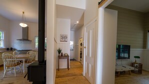 Een keuken met witte kastjes, een houten eettafel en stoelen. Er staat een zwarte kookplaat, een hangende lamp en een schilderij aan de muur.