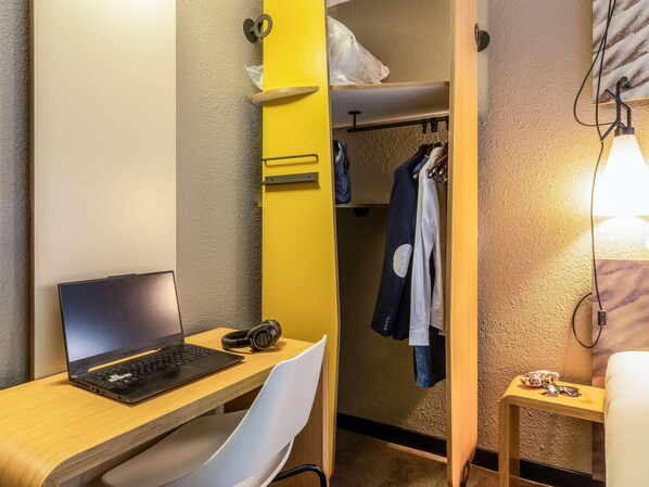 A compact hotel room with a wooden desk, a laptop, headphones, a white chair, a wardrobe with hanging clothes, and a bedside table with a lamp. - Ibis Jouy en Josas Velizy (Jouy-en-Josas)