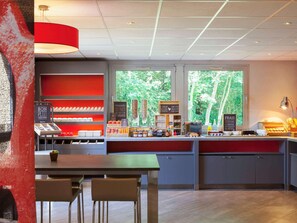 A café with a red counter, shelves with snacks, a coffee machine, and a view of trees through the window. - Ibis Jouy en Josas Velizy (Jouy-en-Josas)