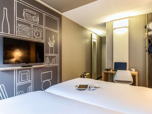 A hotel room with a bed, a TV mounted on the wall, a desk with a laptop, a chair, and a mirror. - Ibis Jouy en Josas Velizy (Jouy-en-Josas)